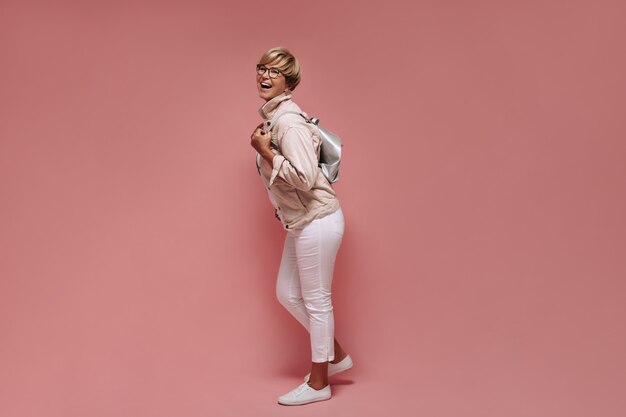 Foto in voller Länge von Frau mit kurzen Haaren und Brille in dünnen hellen Hosen, weißen Turnschuhen und Jacke, die lachen und Tasche auf rosa Hintergrund halten.