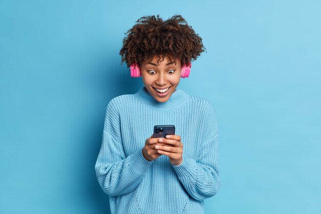 Foto von Afroamerikaner Teenager-Mädchen starrt beeindruckt auf Smartphone-Display liest erstaunliche Nachrichten trägt rosa Stereo-Kopfhörer hört Musik in Strickpullover gekleidet