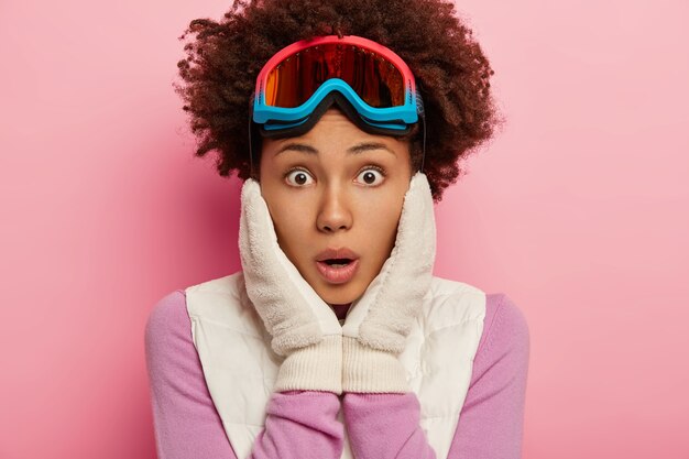 Foto von betäubten emotionalen lockigen weiblichen Blicken mit geschocktem Gesichtsausdruck an der Kamera, hält beide Hände auf Wangen, trägt Skibrille zum Snowboarden, isoliert über rosiger Studiowand