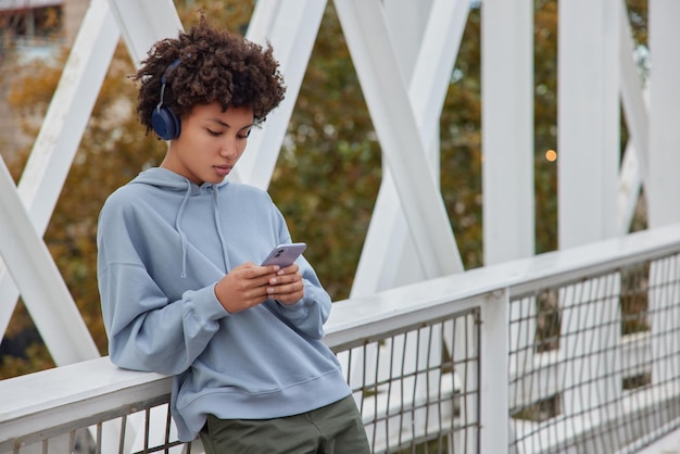 Foto von ernsthaftem lockigem Teenager-Mädchen hört Musik in drahtlosen Kopfhörern hält Handy-Checks Benachrichtigungsposen auf der Brücke im Freien trägt einen lässigen Hoodie, der auf dem Bildschirm des Mobiltelefons konzentriert ist