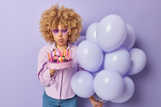 Foto von Frau mit lockigen Haaren bläst Kerzen auf Kuchen hält Haufen aufgeblasener Luftballons trägt Sonnenbrille und Hemd isoliert über violettem Hintergrund Menschen feiern und festliche Anlässe Konzept