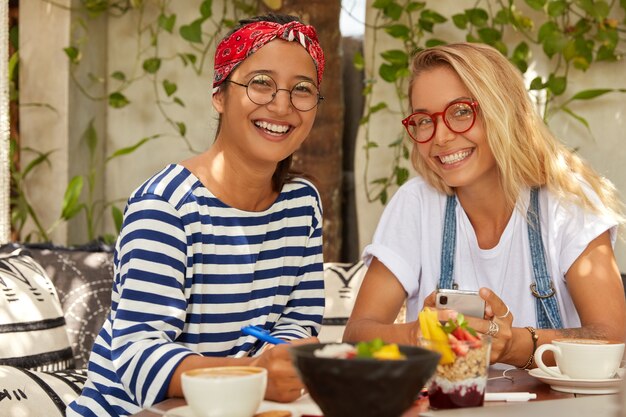 Foto von fröhlichen zwei gemischten Studenten treffen sich im Café, um gemeinsame Aufgaben zu erledigen, leckeres Gericht zu genießen, breit zu lächeln, eine optische Brille zu tragen, auf dem Handy zu plaudern, Übung im Notizblock zu schreiben, Kaffee zu trinken