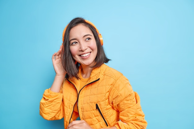 Foto von gut aussehendem Teenager-Mädchen mit östlichem Aussehen hört Musik in modernen Kopfhörern lächelt breit trägt orange modische Jacke isoliert über blauer Wand. Tolle Wiedergabeliste. Musikliebhaber