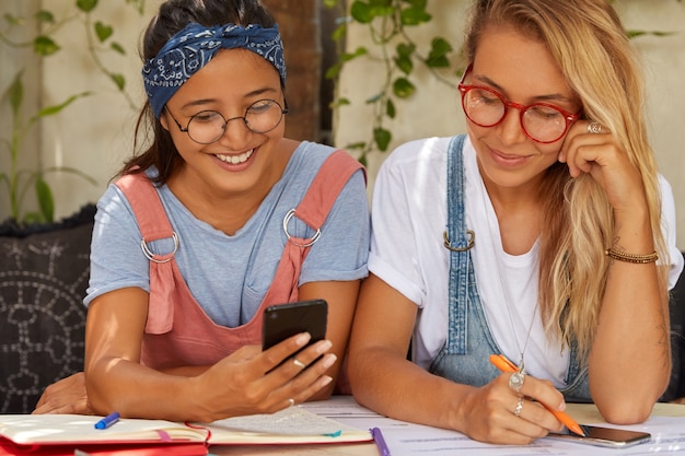 Foto von Schwestern oder Mitarbeitern verwenden Handy, geben sich gegenseitig Ratschläge, übersetzen Artikel, schreiben Aufzeichnungen in Notizblock, posieren auf dem Sofa im Sommergarten tragen optische Brillen, Stirnband, T-Shirt, nutzen Sie kostenloses WLAN