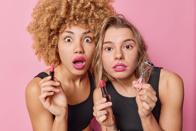 Foto von überraschten jungen Frauen tragen Lippenstift auf, halten kosmetische Produkte, machen Make-up, starren schockiert in die Kamera, wollen schön gekleidet aussehen, lässig isoliert über rosa Hintergrund Schönheitsverfahren