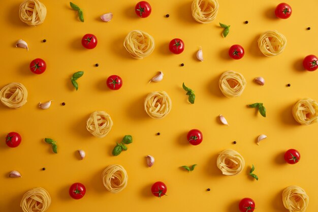 Foto von ungekochten Nudelnestern, die um essbare rote Tomaten, Knoblauch, Pfefferkörner, Basilikum auf gelbem Hintergrund liegen. Kochen nahrhaftes Essen. Italienische traditionelle Küche. Große Produktvielfalt