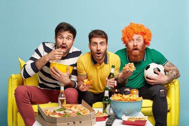 Foto von verblüfften besten männlichen Freunden starren auf den Bildschirm des Fernsehers, halten Bier, essen köstliche Pizza, schockiert mit unerwartetem Ergebnis des Fußballspiels, sitzen auf bequemer gelber Couch, verlorenes Streichholz, isoliert auf Blau