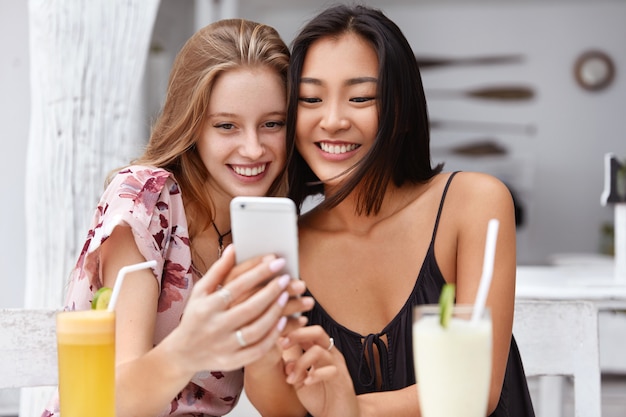 Foto von zufriedenen Frauen gemischter Rassen erhalten gute Nachrichten auf dem Handy, erhalten E-Mails oder machen Selfies mit dem Smartphone, trinken frische Cocktails in der Cafeteria.