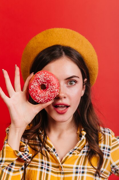 Französische Dame in Baskenmütze und mit rotem Lippenstift, die ihr Gesicht mit rosa Donut bedeckt.