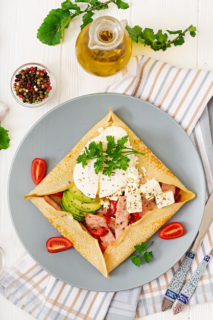 Französische Küche. Frühstück, Mittagessen, Snacks. Pfannkuchen mit pochiertem Ei, Feta-Käse, gebratenem Schinken, Avocado und Tomaten auf weißem Tisch. Draufsicht