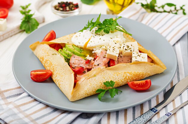 Französische Küche. Frühstück, Mittagessen, Snacks. Pfannkuchen mit pochiertem Ei, Feta-Käse, gebratenem Schinken, Avocado und Tomaten auf weißem Tisch