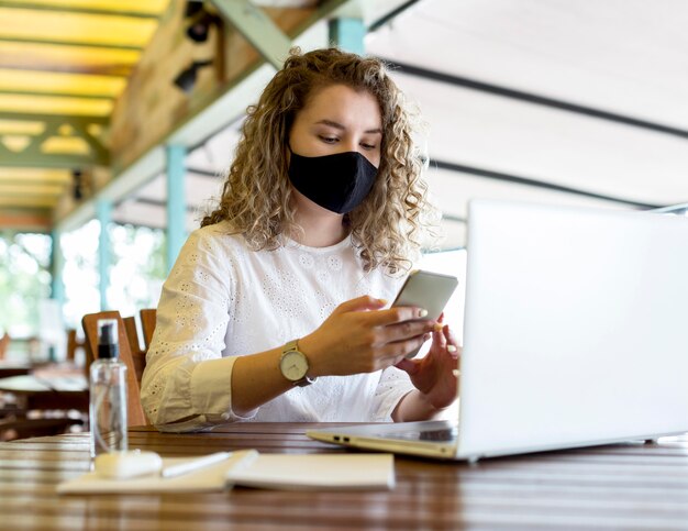 Frau an der Terrasse mit Maske unter Verwendung des Telefons