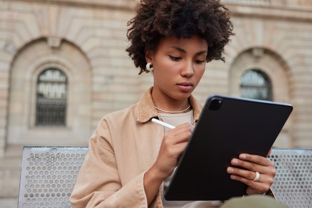 Frau arbeitet am Tablet mit Stylus zeichnet Skizzen für zukünftige Projekte arbeitet an der Bearbeitung von Bildern für die Bildproduktionn sitzt im Freien trägt stilvolle Kleidung