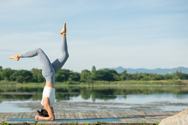 Frau auf einer Yogamatte, zum im Park zu entspannen. Junge sportliche asiatische Frau, die Yoga praktiziert, Kopfstandübung macht, trainiert, Sportbekleidung, Hosen und Top trägt.