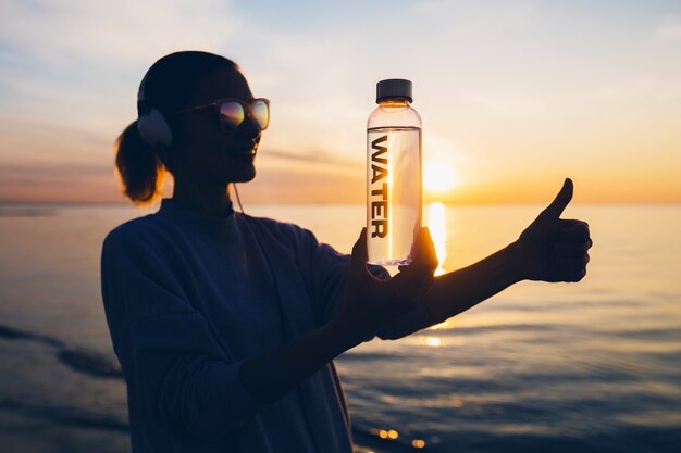 Frau bei Sonnenaufgang am Strand, die eine Flasche Wasser hält und eine positive Geste zeigt, Daumen hoch