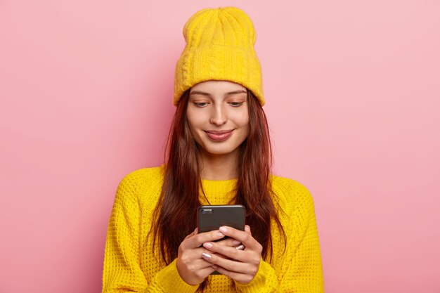 Frau benutzt ein Smartphone, hat langes glattes Haar, trägt gelben Pullover und Hut, hat zufriedenen Gesichtsausdruck, posiert vor rosa Hintergrund