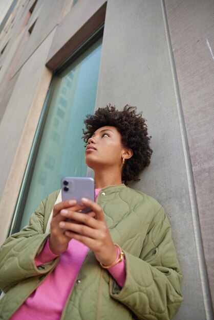 Frau benutzt Telefongespräche mit Fried hält Handy, das irgendwo fokussiert ist, trägt lässige Jackenposen in der Nähe der Wand des modernen Stadtgebäudes