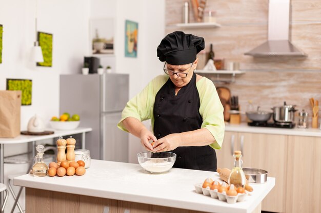 Frau bricht Ei über Mehl und macht Teig für Backwaren. Älterer Konditor, der Ei auf Glasschüssel für Kuchenrezept in der Küche knackt, von Hand mischt, Zutaten knetet, um hausgemachten Kuchen zuzubereiten