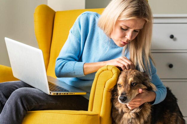 Frau, die am Laptop vom Sessel während der Pandemie arbeitet und ihren Hund streichelt