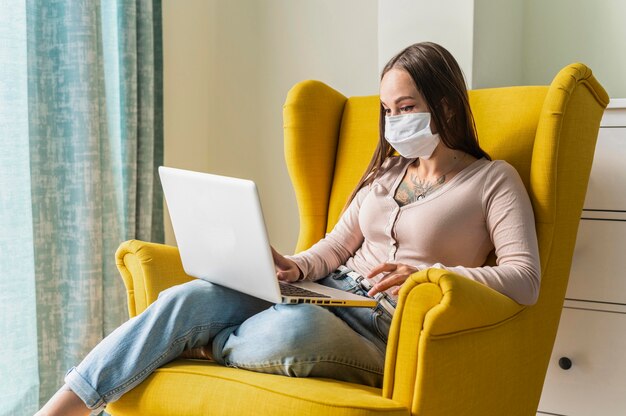 Frau, die am Laptop vom Sessel während der Pandemie beim Tragen der medizinischen Maske arbeitet