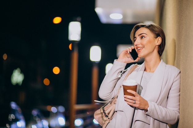 Frau, die am Telefon spricht und Kaffee draußen in der Straße nachts trinkt