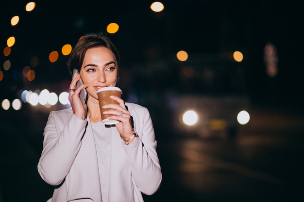 Frau, die am Telefon spricht und Kaffee draußen in der Straße nachts trinkt