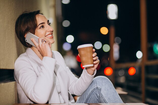 Frau, die am Telefon spricht und Kaffee draußen in der Straße nachts trinkt