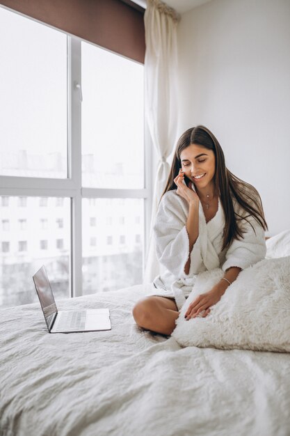 Frau, die an Computer im Bett arbeitet und am Telefon spricht