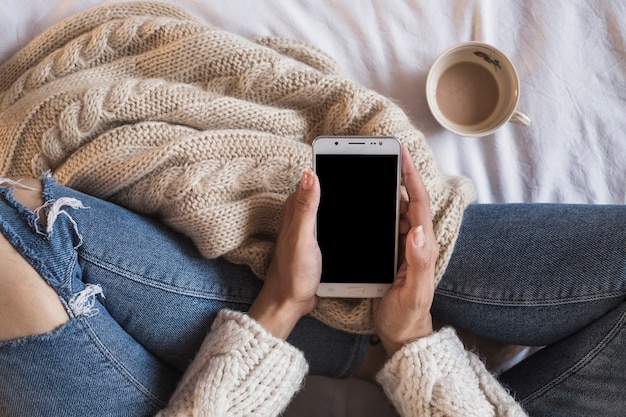 Frau, die auf Bett mit Telefon und Tasse Kaffee sitzt