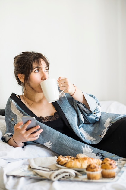 Kostenloses Foto frau, die auf betttrinkbecher kaffee sich lehnt