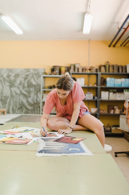 Frau, die auf dem Werktisch tut Malerei auf Segeltuchpapier in der Werkstatt sitzt