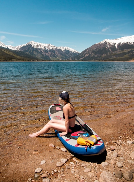 Frau, die auf einem Paddelboard nahe dem Wasser sitzt und die Aussicht auf einen schneebedeckten Berg und einen klaren Himmel genießt