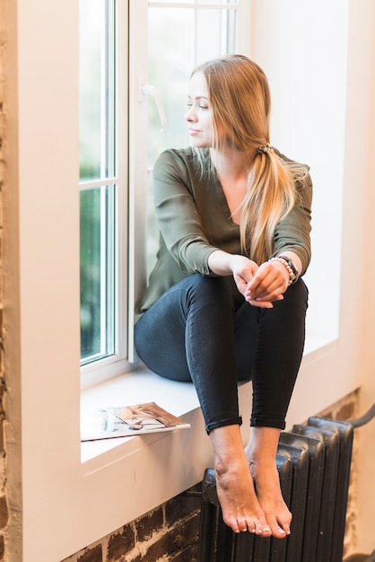 Frau, die auf Fensterbrett mit ihrem Bein auf dem Kühler weg schaut sitzt