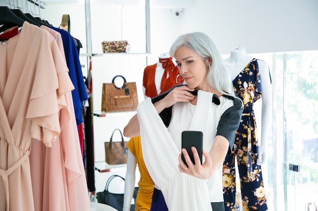 Frau, die auf smartphone im modegeschäft spricht und kleid zeigt. mittlerer schuss. boutique-kunden- oder kommunikationskonzept