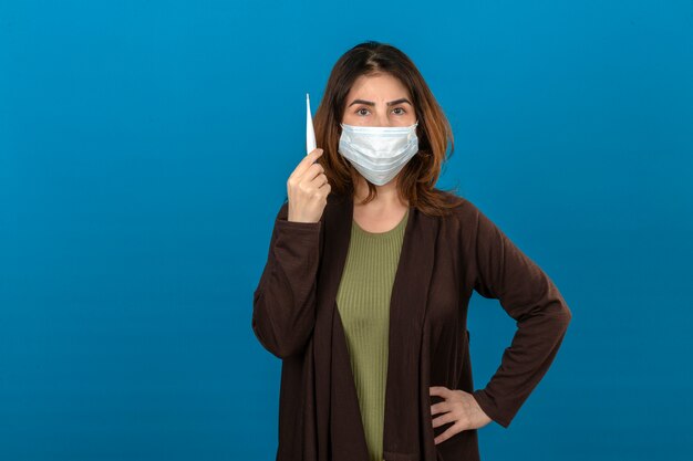 Frau, die braune Strickjacke in der medizinischen Schutzmaske trägt, die digitales Thermometer in der Hand mit ernstem Gesicht über isolierter blauer Wand hält