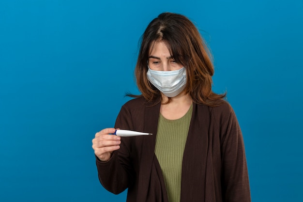 Frau, die braune Strickjacke in der medizinischen Schutzmaske trägt, die krank hält digitales Thermometer hält, das es mit traurigem Ausdruck auf Gesicht über isolierter blauer Wand betrachtet