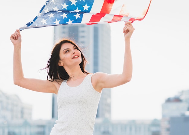 Frau, die breite amerikanische Flagge auf Hintergrund des Geschäftszentrums wellenartig bewegt