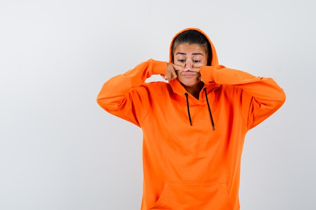 Frau, die die Nase mit den Fingern im orangefarbenen Hoodie berührt und lustig aussieht