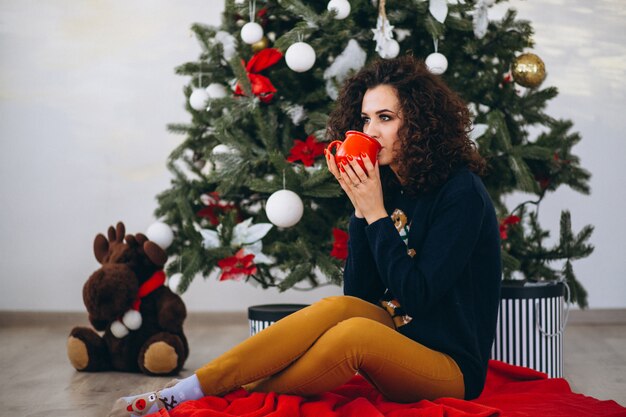 Frau, die durch Weihnachtsbaum sitzt und Tee trinkt