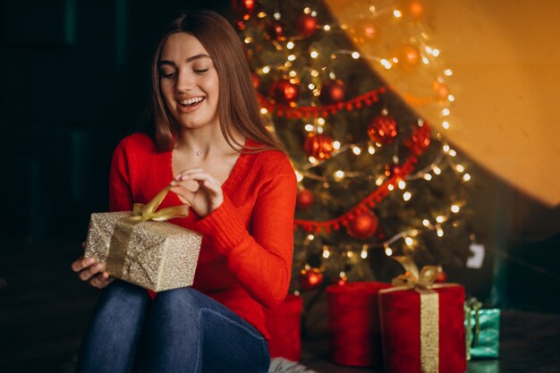Frau, die durch Weihnachtsbaum sitzt und Weihnachtsgeschenk auspackt