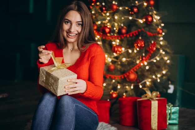 Frau, die durch Weihnachtsbaum sitzt und Weihnachtsgeschenk auspackt