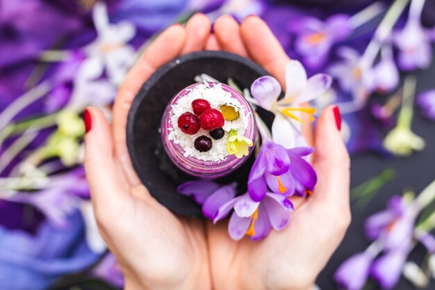 Frau, die ein Glas des veganen Smoothie hält, der mit Beeren gekrönt wird, umgeben von lila Frühlingsblumen