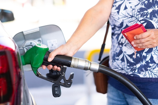 Frau, die eine benzinpumpe hält, um ihr silbernes auto an der tankstelle zu tanken, und eine brieftasche hält, um zu bezahlen