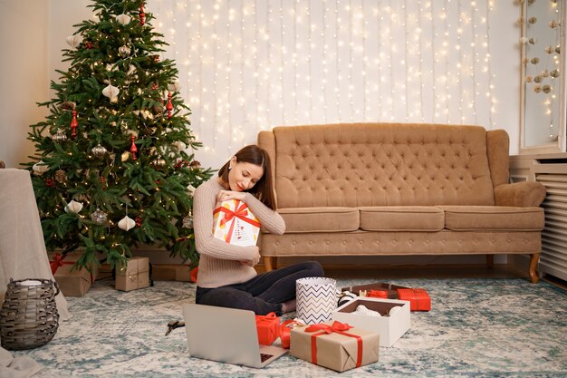 Frau, die eine Geschenkbox umarmt, während sie zwischen vielen Geschenken im weihnachtlich geschmückten Leben sitzt