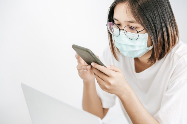 Frau, die eine Hygienemaske trägt, ein Smartphone spielt und einen Laptop hat.