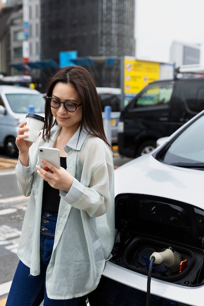 Frau, die eine Kaffeepause macht, während ihr Elektroauto auflädt und Smartphone verwendet
