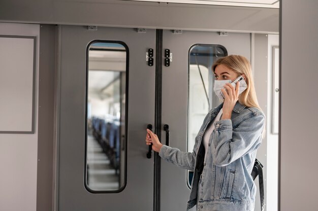 Frau, die eine medizinische Maske trägt und telefoniert, während sie sich auf die Reise mit dem Zug vorbereitet