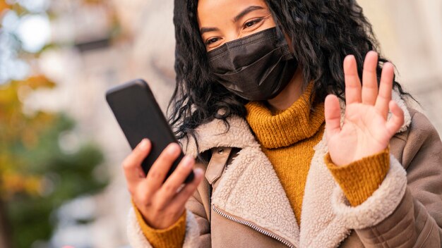 Frau, die eine medizinische Maske trägt, während sie einen Videoanruf auf ihrem Smartphone hat