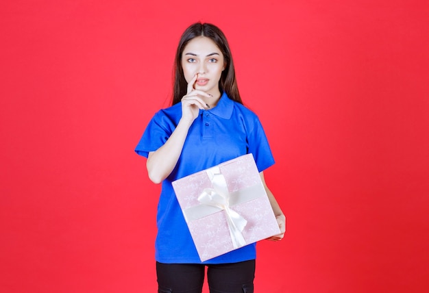 Frau, die eine rosa Geschenkbox mit weißem Band hält und verwirrt aussieht oder eine gute Idee hat.