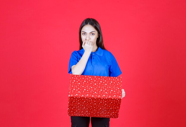 Frau, die eine rote Geschenkbox mit weißen Punkten hält und verwirrt und zögerlich aussieht.